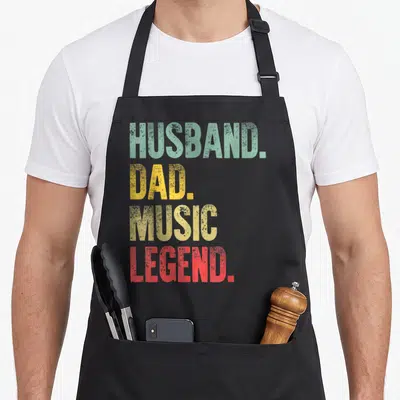 Music legend husband apron - Stacked HUSBAND DAD MUSIC LEGEND text in green yellow gold and red distressed font — shown on a black cotton canvas apron against a white background