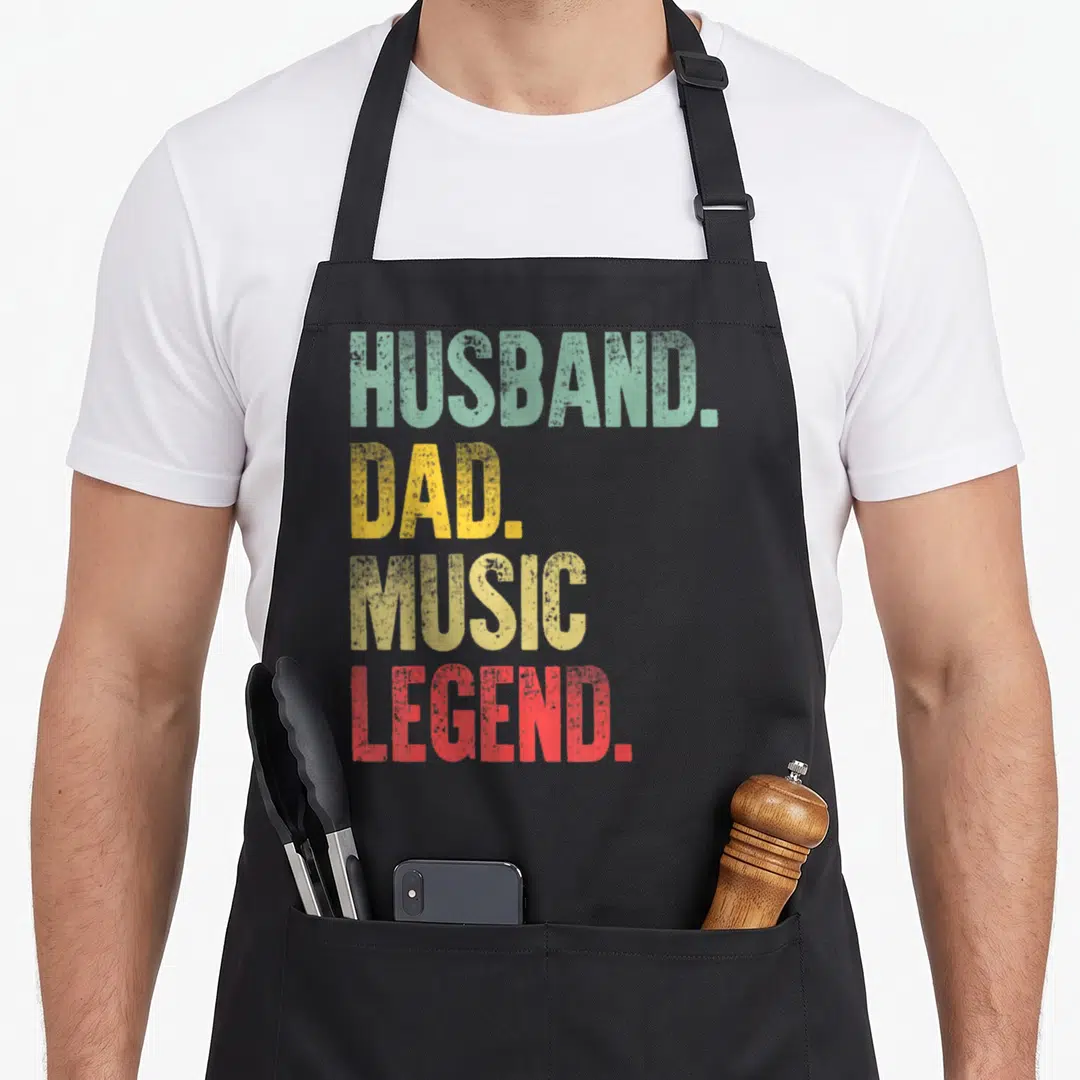 Music legend husband apron - Stacked HUSBAND DAD MUSIC LEGEND text in green yellow gold and red distressed font — shown on a black cotton canvas apron against a white background