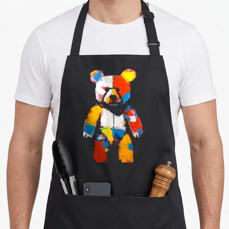 Motif bear apron - Multicolored teddy bear in red blue yellow and white cartoon style on black background — shown on a black cotton canvas apron against a white background