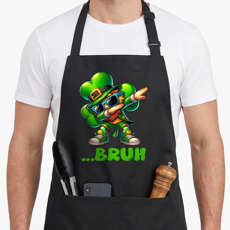Bruh st patrick day aprons - Cartoon leprechaun dabbing in front of a green shamrock with ...BRUH text below — shown on a black cotton canvas apron against a white background
