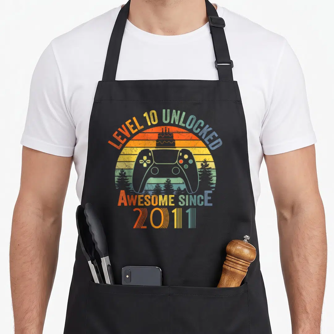 Unlocked video birthday apron - Retro sun with grey game controller cake and LEVEL 10 UNLOCKED text in orange yellow — shown on a black cotton canvas apron against a white background