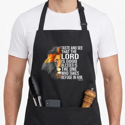 Faith god christian apron - Lion head split vertically with stacked white text reading Taste and See that the Lord is — shown on a black cotton canvas apron against a white background
