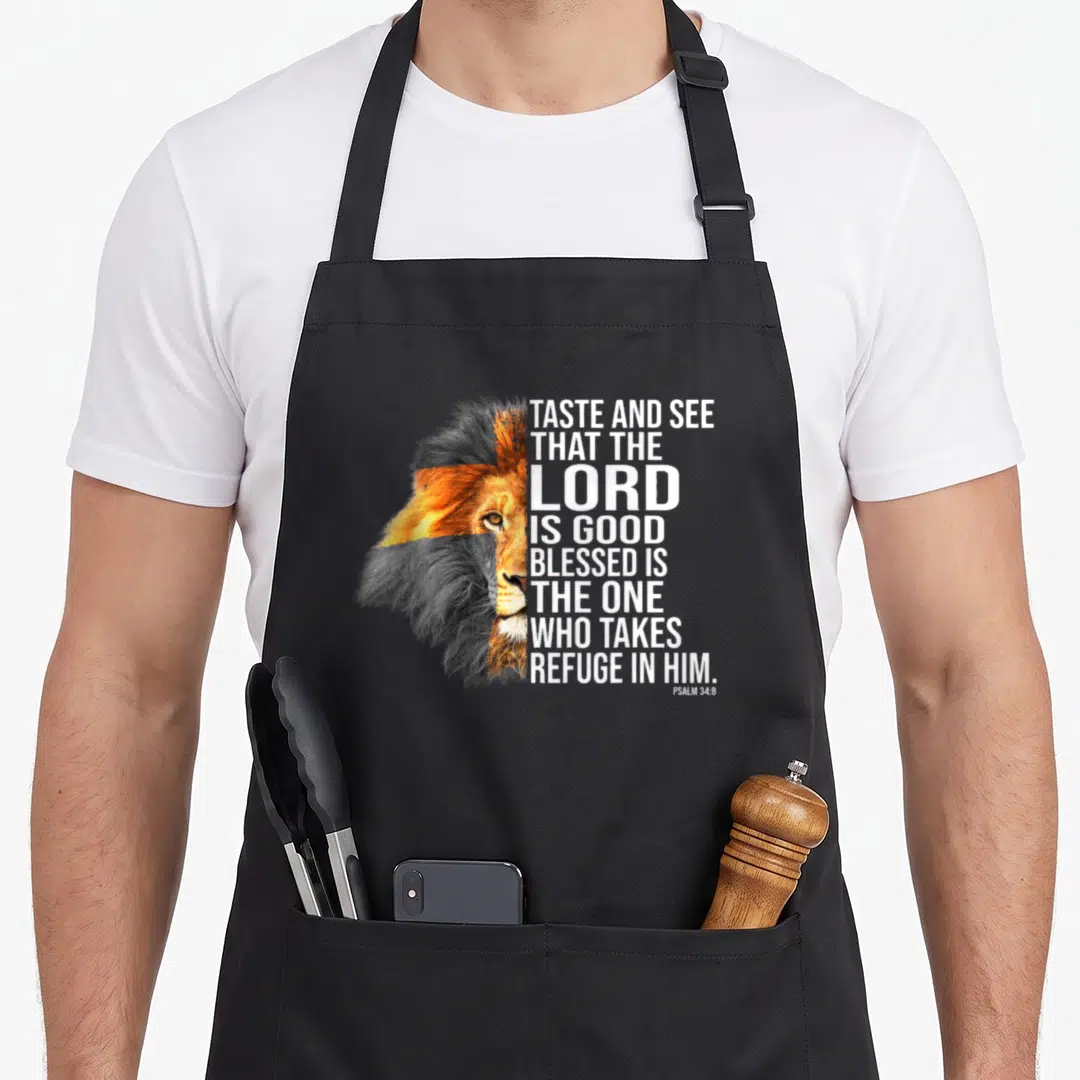 Faith god christian apron - Lion head split vertically with stacked white text reading Taste and See that the Lord is — shown on a black cotton canvas apron against a white background