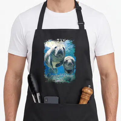 Crystal florida apron - Two manatees swimming in blue water with Crystal River Florida text below — shown on a black cotton canvas apron against a white background