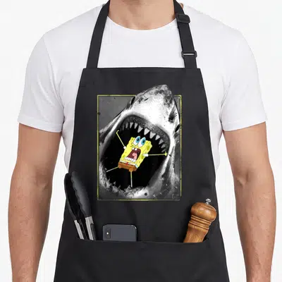 Squarepant funny hustler apron - Cartoon yellow character floating inside open grey shark mouth with yellow border — shown on a black cotton canvas apron against a white background