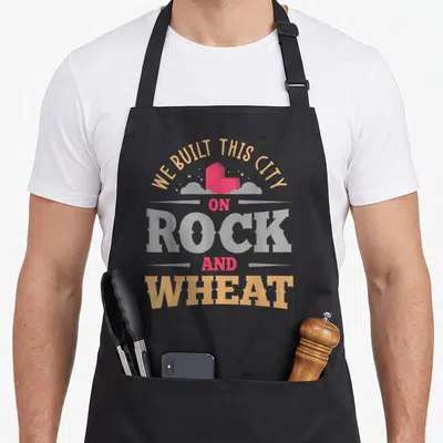 Built apron or apron - Stacked text We Built This City On Rock And Wheat with red brick and grey clouds — shown on a black cotton canvas apron against a white background