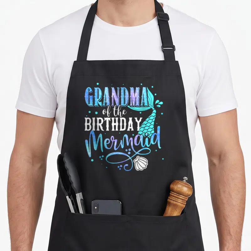 Birthday mermaid grandma in apron - Stacked text GRANDMA of the BIRTHDAY Mermaid with blue purple teal mermaid tail — shown on a black cotton canvas apron against a white background