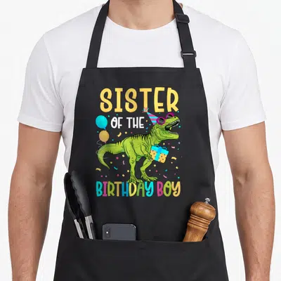 Squad dinosaur apron - Green T-Rex with party hat holding gift under Sister of the Birthday Boy text — shown on a black cotton canvas apron against a white background