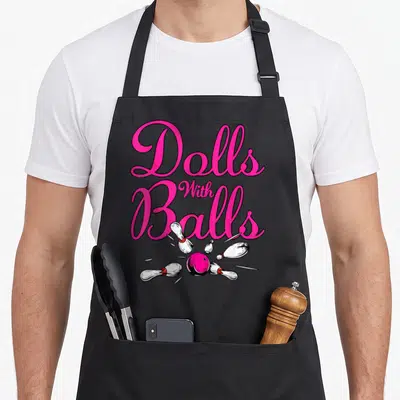 Ball doll apron - Pink script text reading Dolls With Balls above pink bowling ball scattering five white pins — shown on a black cotton canvas apron against a white background