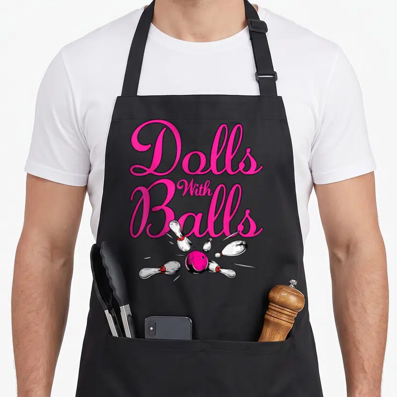 Ball doll apron - Pink script text reading Dolls With Balls above pink bowling ball scattering five white pins — shown on a black cotton canvas apron against a white background