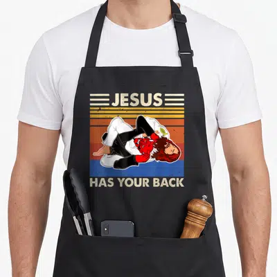 Jesus jitsu apron - Cartoon Jesus in gi holding devil over striped background with text Jesus Has Your Back — shown on a black cotton canvas apron against a white background