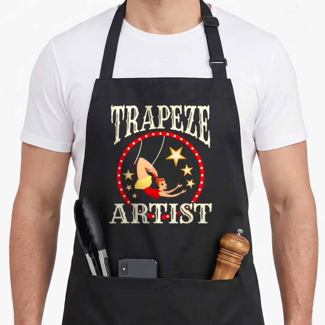Trapeze circus apron - Cartoon trapeze artist hanging from a bar inside a red ring with stars and cream text — shown on a black cotton canvas apron against a white background