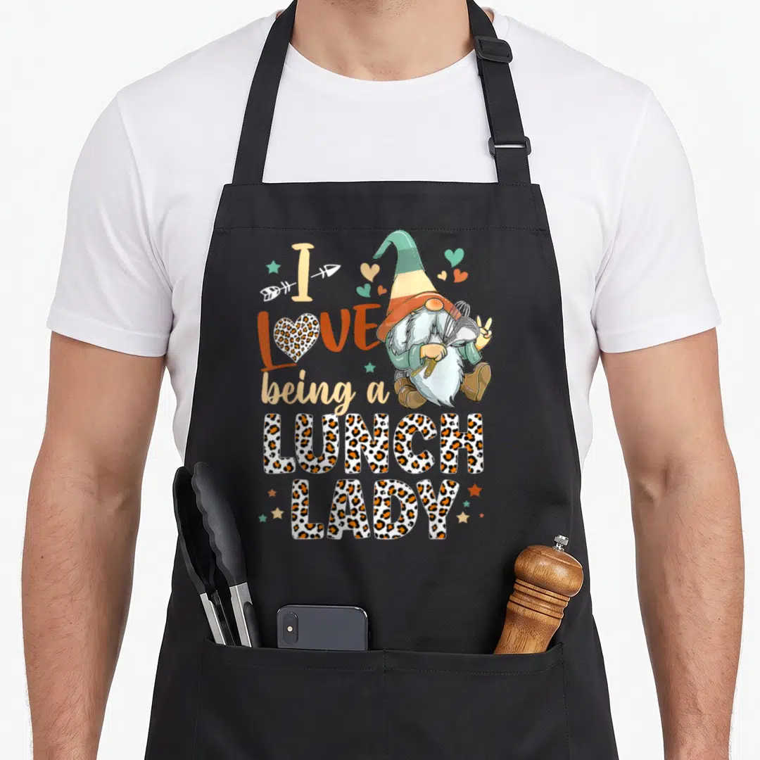 Gnome lunch lady apron - Gnome holding whisk next to I LOVE being a LUNCH LADY text in leopard print — shown on a black cotton canvas apron against a white background