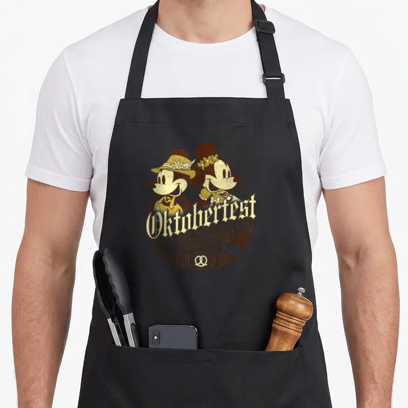 Friend oktoberfest apron - Mickey and Minnie in brown and gold Oktoberfest badge with pretzels and German text — shown on a black cotton canvas apron against a white background