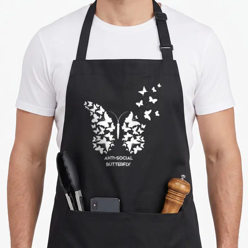 Funny antisocial butterfly apron - White text ANTI-SOCIAL BUTTERFLY below a large butterfly shape made of many smaller — shown on a black cotton canvas apron against a white background