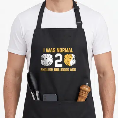Normal english apron - Two cartoon bulldog heads flanking a white number 2 with yellow text above and below — shown on a black cotton canvas apron against a white background