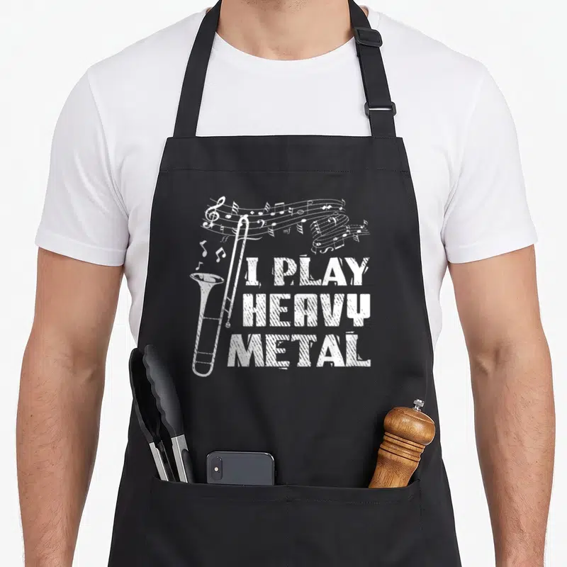 Trombone heavy metal apron - White distressed trombone with musical staff and stacked text I PLAY HEAVY METAL — shown on a black cotton canvas apron against a white background