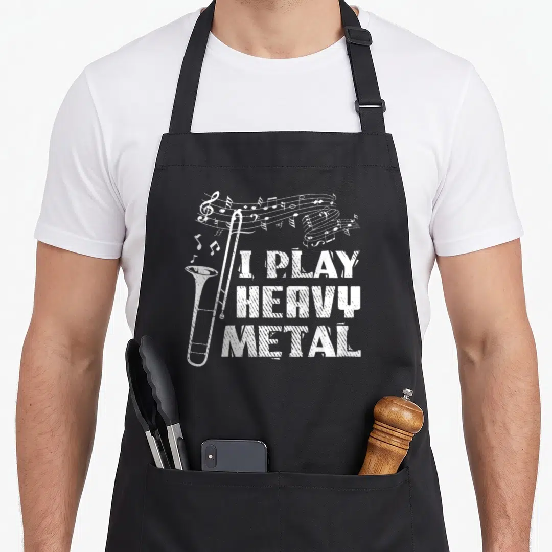 Trombone heavy metal apron - White distressed trombone with musical staff and stacked text I PLAY HEAVY METAL — shown on a black cotton canvas apron against a white background