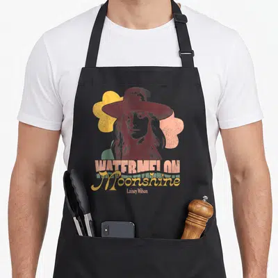 Lainey watermelon apron - Retro portrait of woman in hat with WATERMELON Moonshine Lainey Wilson text and colorful — shown on a black cotton canvas apron against a white background
