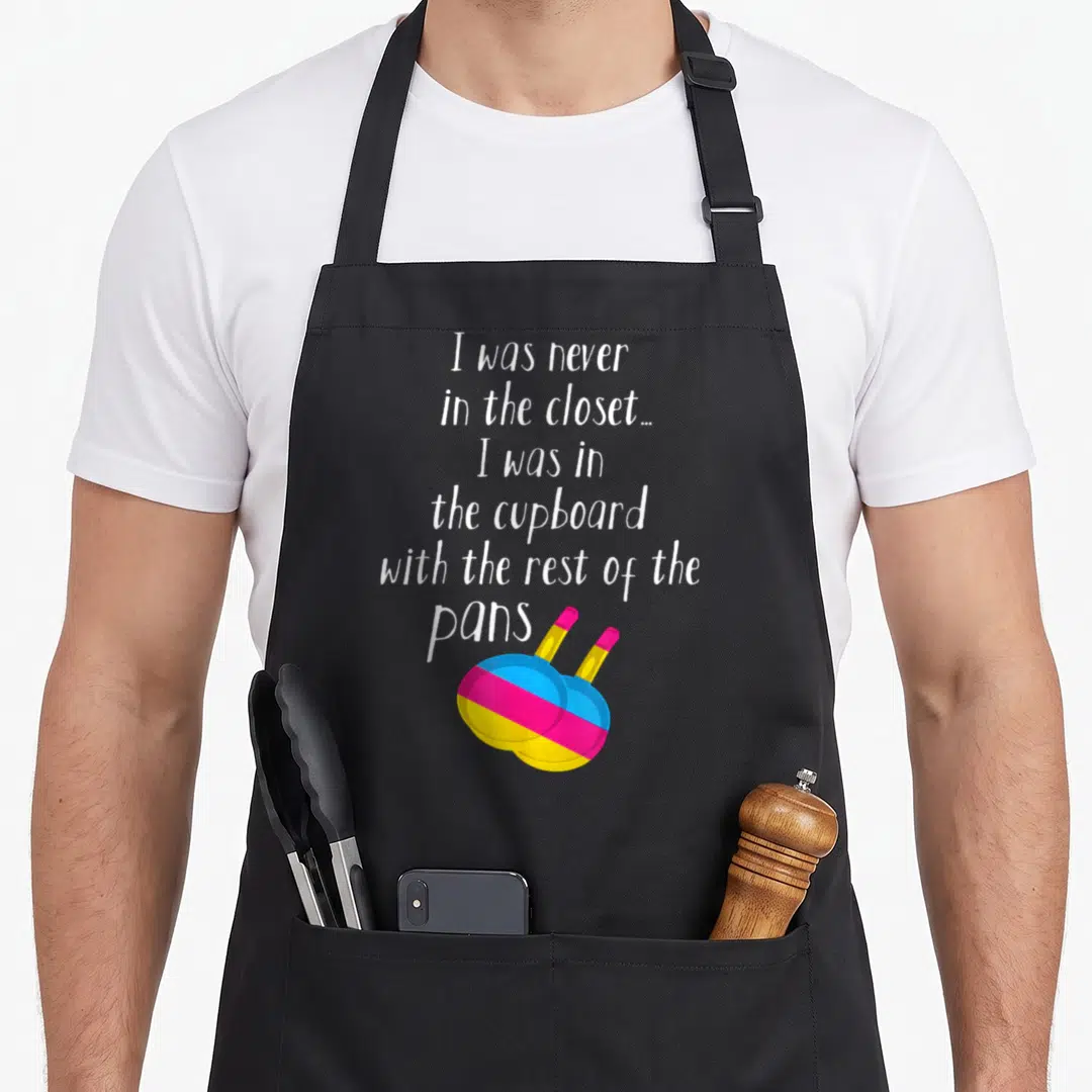 Pansexual meme apron - White text above two overlapping frying pans in blue pink and yellow stripes — shown on a black cotton canvas apron against a white background