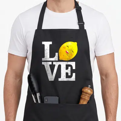 Fresh lemonade apron - White distressed LOVE text with a smiling yellow lemon character replacing the letter O — shown on a black cotton canvas apron against a white background
