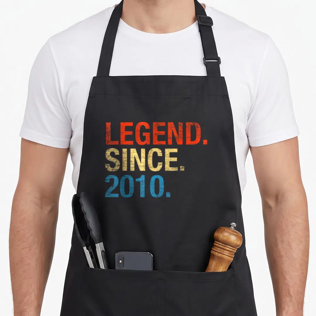 Legend 12th birthday apron - Stacked red yellow blue distressed text reading LEGEND SINCE 2010 on black fabric — shown on a black cotton canvas apron against a white background