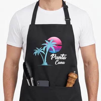 Dainty punta cana apron - Two blue palm trees on pink purple white gradient sun with Punta Cana text — shown on a black cotton canvas apron against a white background