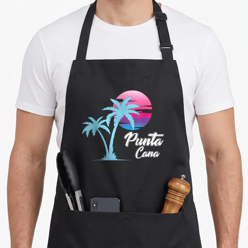 Dainty punta cana apron - Two blue palm trees on pink purple white gradient sun with Punta Cana text — shown on a black cotton canvas apron against a white background