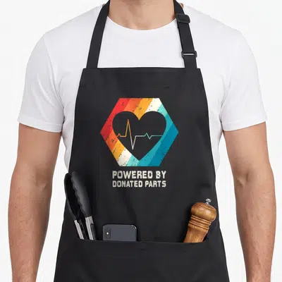 Recipient survivor organ apron - Grey heart with heartbeat line inside striped hexagon and stacked text below — shown on a black cotton canvas apron against a white background