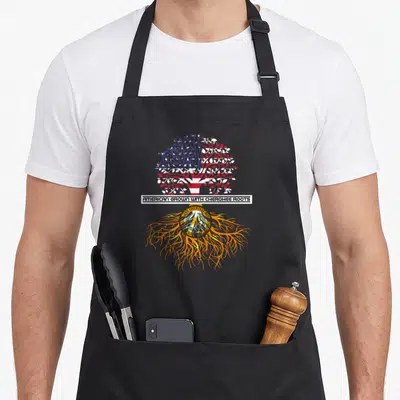 Grown native american apron - Tree with flag canopy, orange roots, circular seal, and text bar AMERICAN GROWN — shown on a black cotton canvas apron against a white background