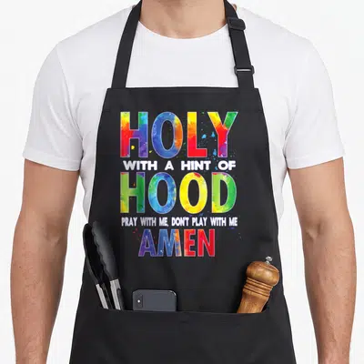 Holy amen apron - Stacked colorful watercolor text reading HOLY WITH A HINT OF HOOD PRAY WITH ME AMEN — shown on a black cotton canvas apron against a white background