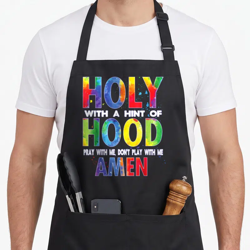 Holy amen apron - Stacked colorful watercolor text reading HOLY WITH A HINT OF HOOD PRAY WITH ME AMEN — shown on a black cotton canvas apron against a white background