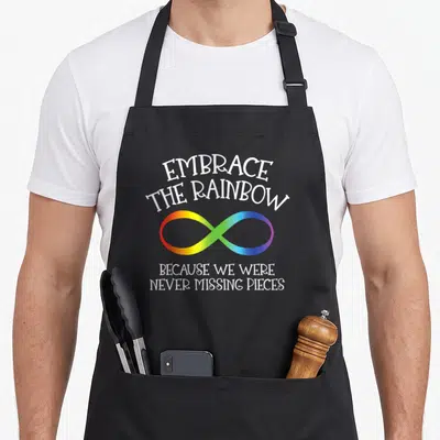 Awareness rainbow apron - White text reading EMBRACE THE RAINBECAUSE WE WERE NEVER MISSING PIECES above and below — shown on a black cotton canvas apron against a white background