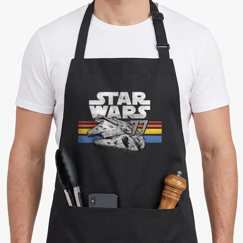 Line falcon apron - Star Wars 77 retro text over Millennium Falcon and four colored stripes — shown on a black cotton canvas apron against a white background