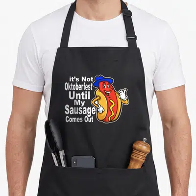 Meme sausage apron - Cartoon hot dog with blue hat next to stacked white text reading it's Not Oktoberfest Until My — shown on a black cotton canvas apron against a white background