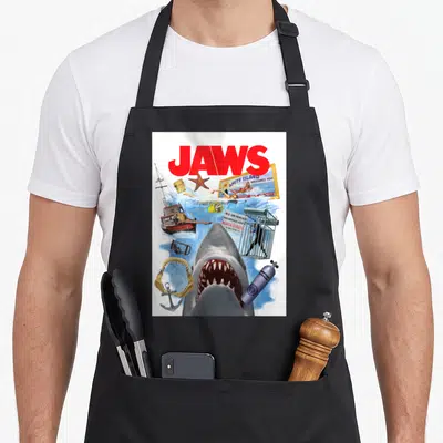 50th kitchen island apron - Red JAWS text, grey shark head, Amity Island sign, diving cage, and boat collage — shown on a black cotton canvas apron against a white background