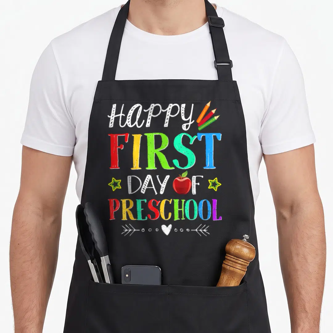 Teacher student preschool apron - Stacked happy first day of preschool text with red apple and three colored pencils — shown on a black cotton canvas apron against a white background