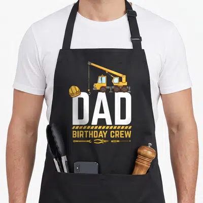 Birthday crew construction apron - Cartoon crane, hard hat, DAD text, striped border, wrench pliers screwdriver — shown on a black cotton canvas apron against a white background