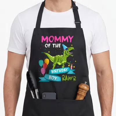 Birthday rex dinosaur apron - MOMMY OF THE BIRTHDAY BOY RAWR text with green T-Rex wearing party hat and sunglasses — shown on a black cotton canvas apron against a white background