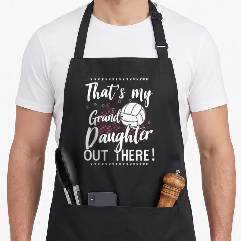 Player grandma granddaughter aprons - That's my Grand Daughter Out There text around volleyball with hearts and stars — shown on a black cotton canvas apron against a white background