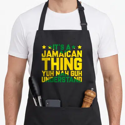 Jamaican island pride apron - Stacked green and yellow text reading IT'S A JAMAICAN THING YUH NAH GUH UNDERSTAND — shown on a black cotton canvas apron against a white background