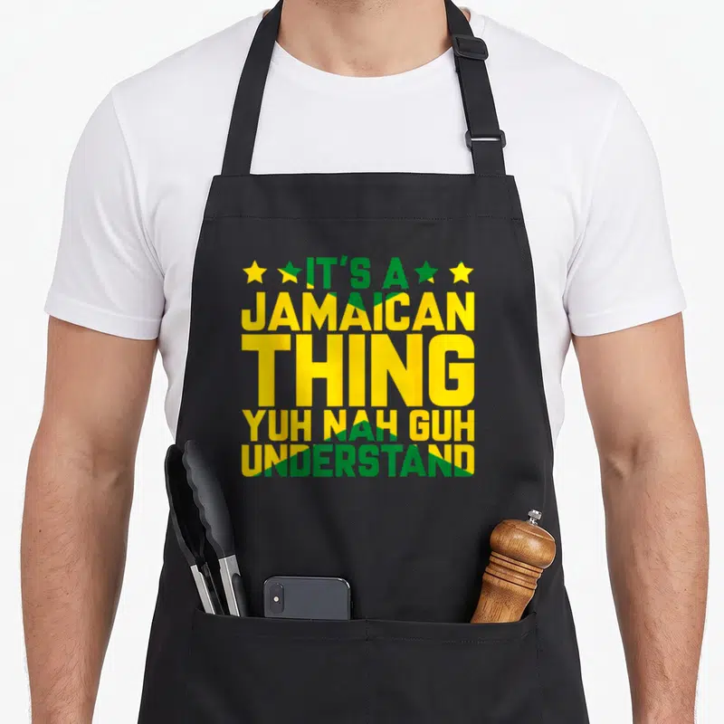 Jamaican island pride apron - Stacked green and yellow text reading IT'S A JAMAICAN THING YUH NAH GUH UNDERSTAND — shown on a black cotton canvas apron against a white background