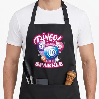 Night bingo aprons - Four numbered bingo balls with BINGO text and white sparkles in magenta and blue — shown on a black cotton canvas apron against a white background
