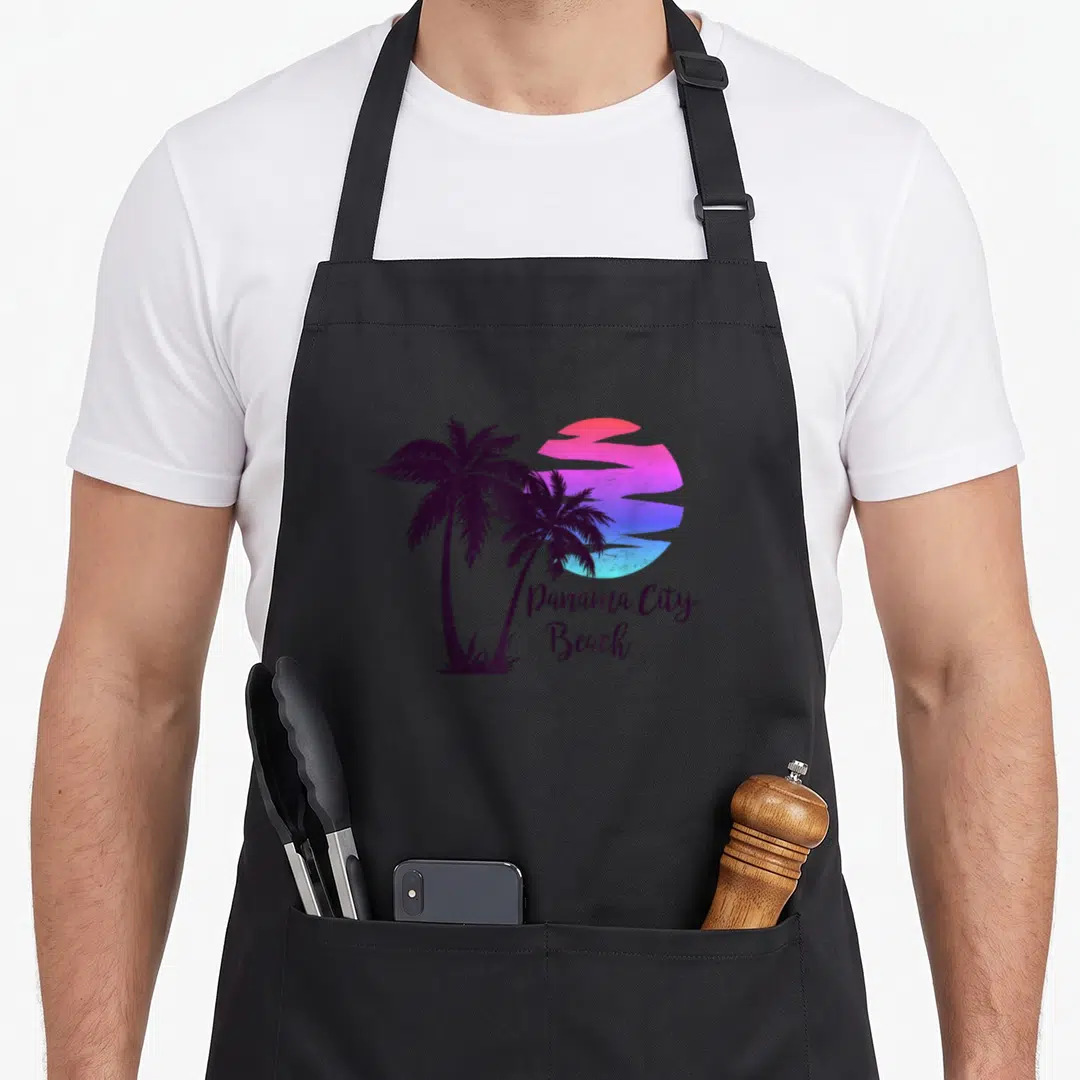 Panama spring aprons - Two purple palm trees beside a pink blue gradient sun with Panama City Beach script text — shown on a black cotton canvas apron against a white background