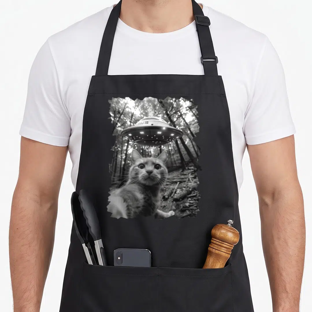 Ufo alien funny cat apron - Black and white photo of a cat reaching a paw forward with a UFO hovering above in a forest — shown on a black cotton canvas apron against a white background