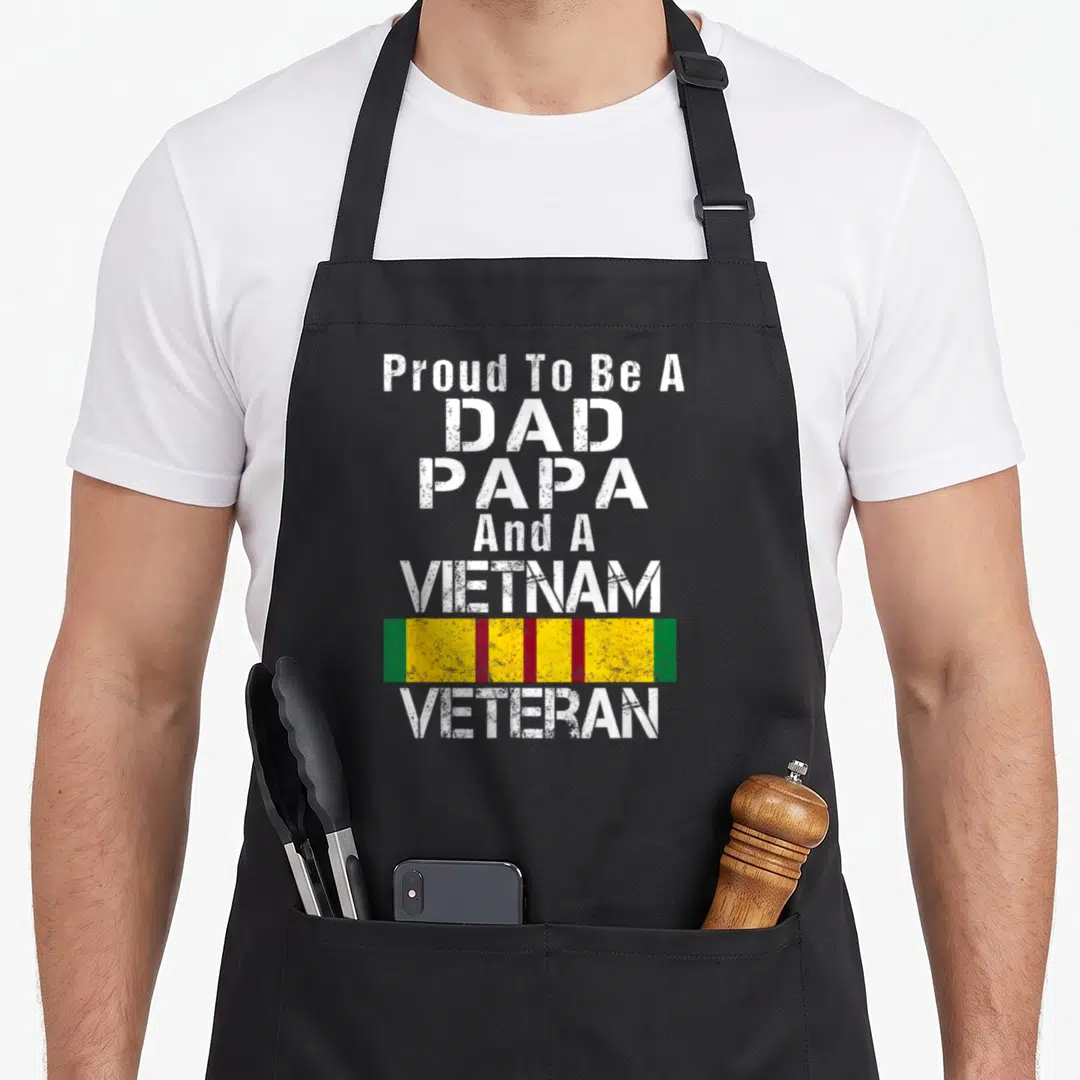 Proud papa apron - Distressed white text Proud To Be A DAD PAPA And A VIETNAM VETERAN with colored ribbon graphic — shown on a black cotton canvas apron against a white background