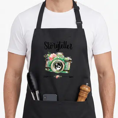Storyteller photographer apron - Watercolor camera with pink flowers and arrow beneath black Storyteller script — shown on a black cotton canvas apron against a white background