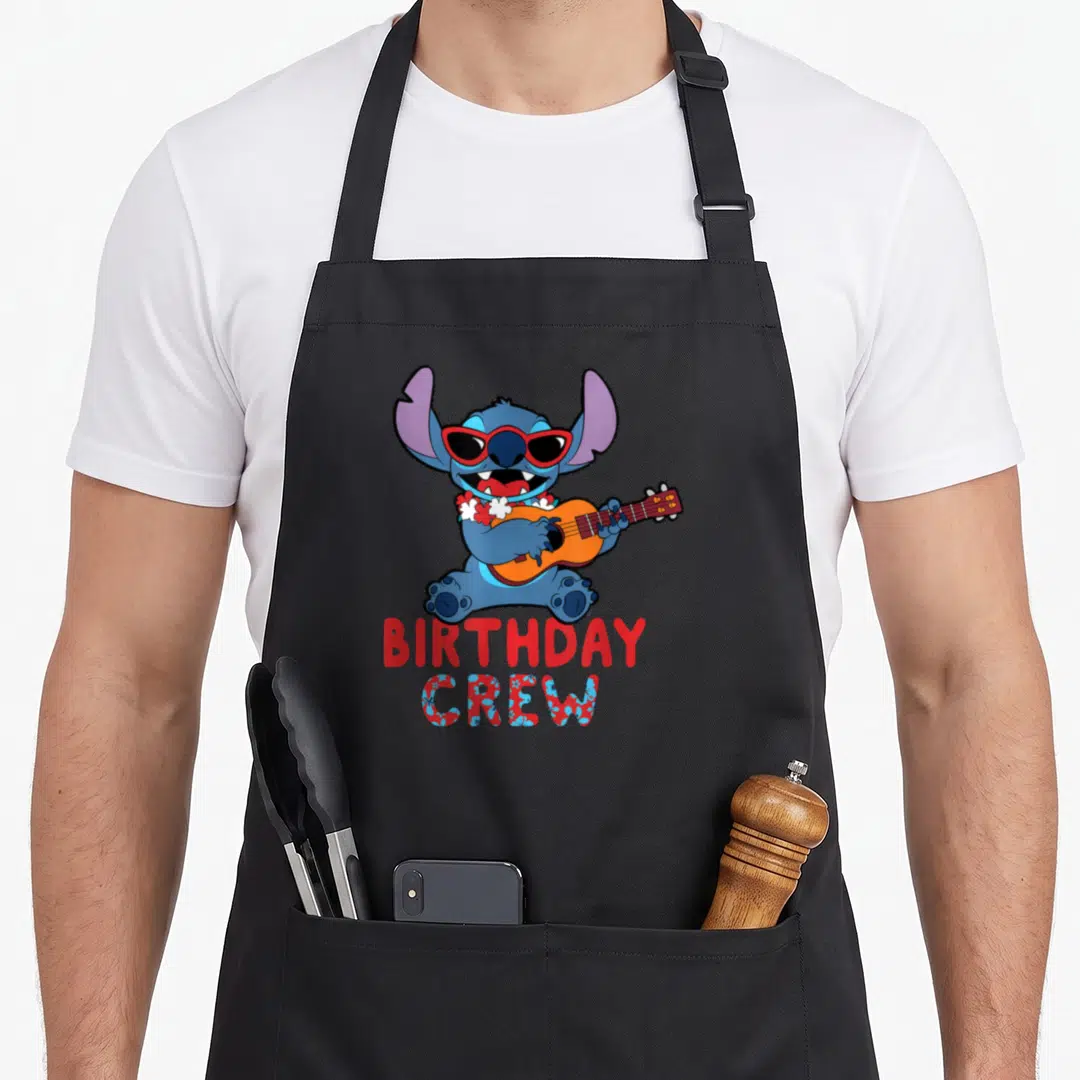 Tropical crew birthday apron - Blue character with red sunglasses holding orange ukulele above BIRTHDAY CREW floral text — shown on a black cotton canvas apron against a white background