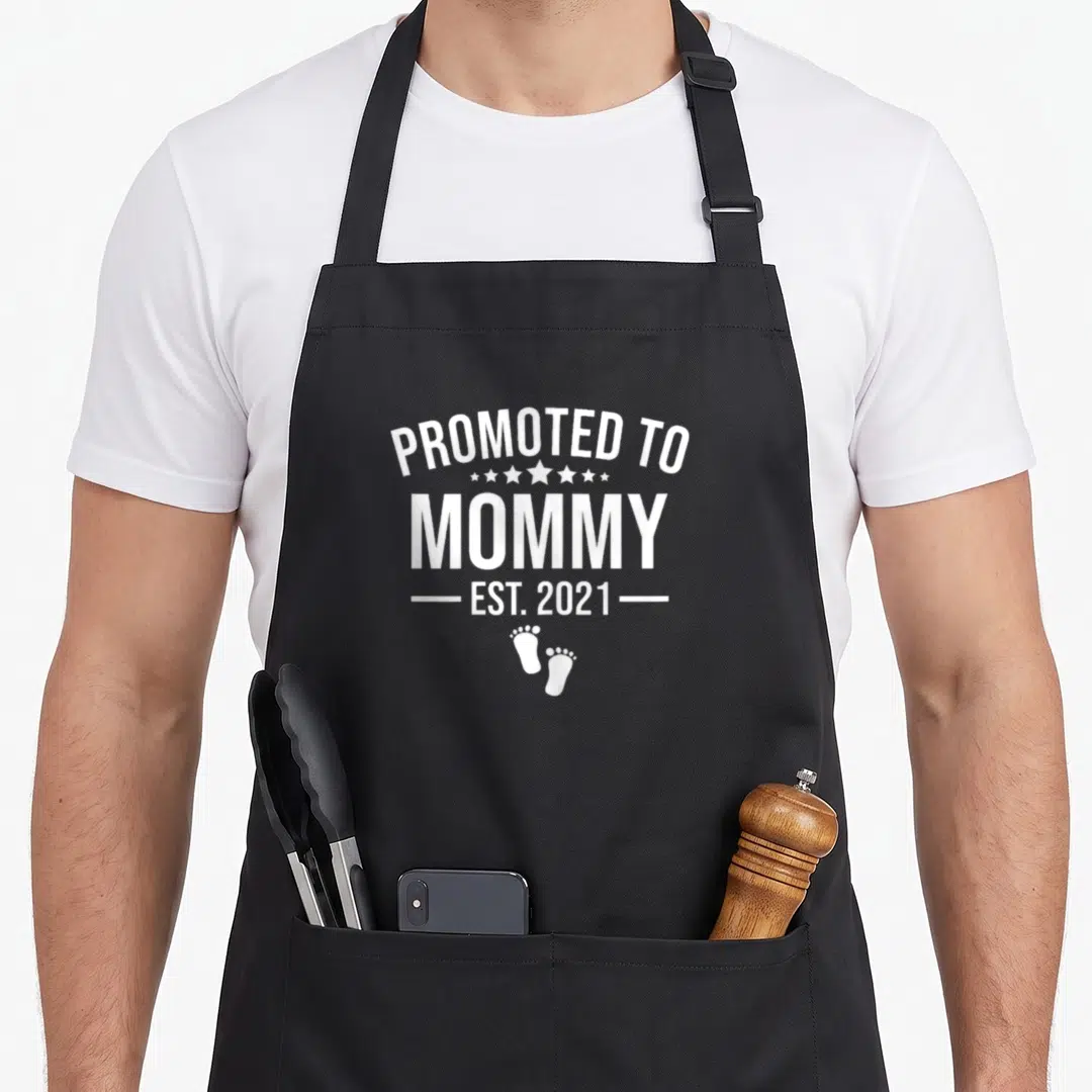 Promoted mother's apron - White PROMOTED TO MOMMY EST 2021 text with stars and two baby footprints — shown on a black cotton canvas apron against a white background