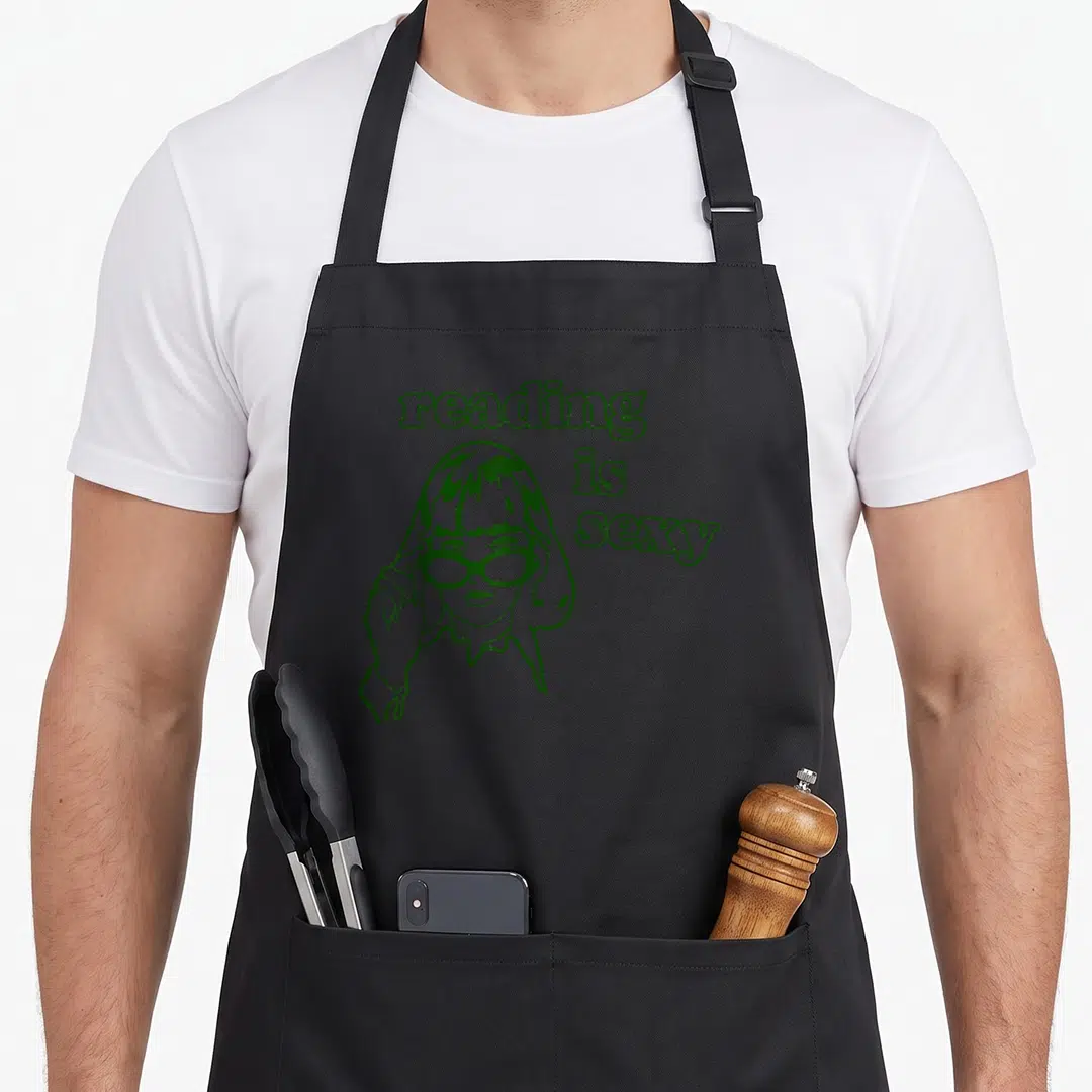 Rory bookworm apron - green line art woman with glasses and stacked text reading issexy — shown on a black cotton canvas apron against a white background